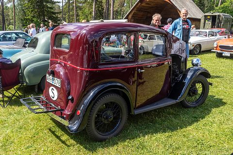 The rear quarter of a maroon saloon with a luggage grid and a spare wheel, parked on grass among cars. Opens a larger view