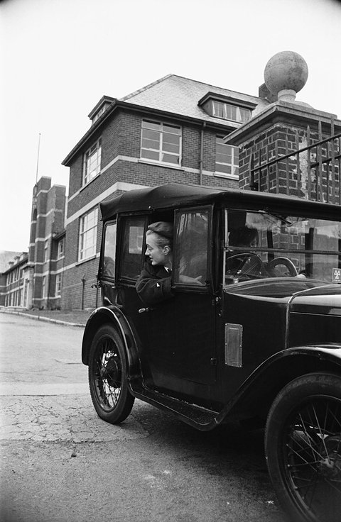 Black and white photograph of a dark upright saloon in a street, a tall building behind it. Opens a larger view
