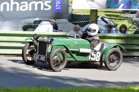 A green racing two-seater with the number 58 driven by a helmeted driver past spectators behind a barrier. Opens a larger view