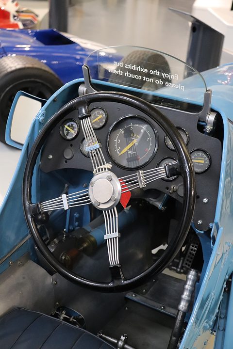 Looking down into a narrow cockpit at a four-spoke steering wheel and two dials. Opens a larger view