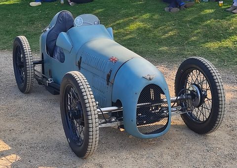 A pale blue single-seat racer with wire wheels and a cowled radiator on gravel. Opens a larger view