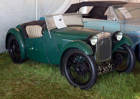 A dark green two-seater with a cowled radiator, cream seats and wire wheels on grass in a tent. Opens a larger view