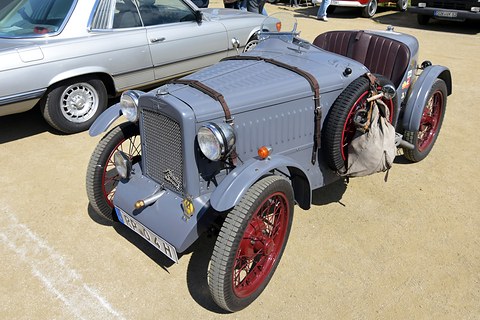 A grey two-seat special with a louvred bonnet, leather straps and red wire wheels on sand. Opens a larger view