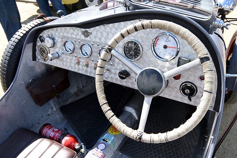 An engine-turned aluminium dashboard with many dials behind a cream steering wheel. Opens a larger view