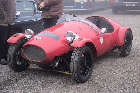 A red rounded two-seat special with a grille and wire wheels on a gravel yard, people behind. Opens a larger view