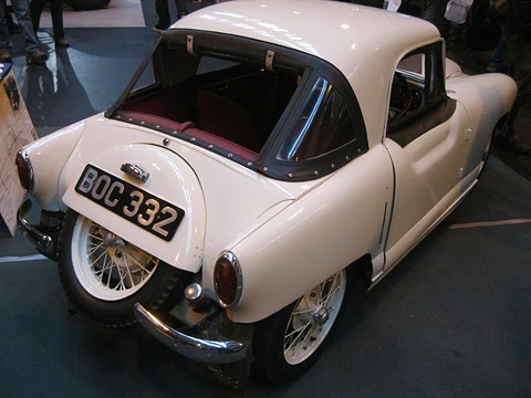 The rear of a cream rounded coupé with a spare wheel on its tail and wire wheels in a dark hall. Opens a larger view
