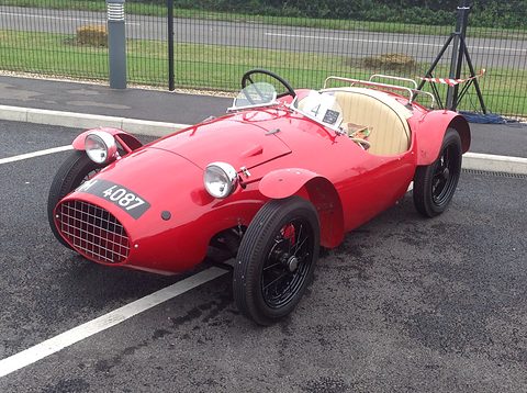 A red rounded two-seat special with a mesh grille and wire wheels in a car park. Opens a larger view
