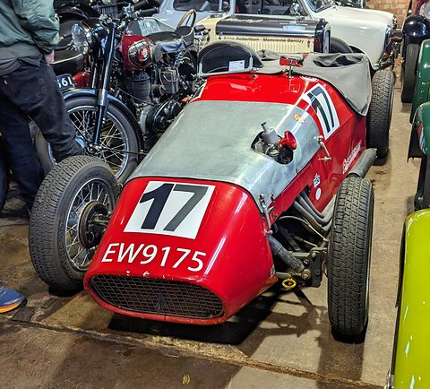 A red single-seat style special with an aluminium bonnet, racing number 17 and wire wheels among motorcycles. Opens a larger view
