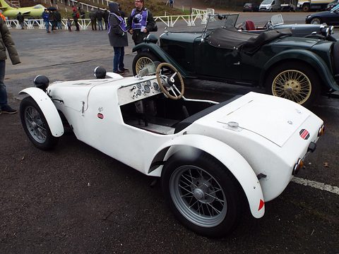 A white open two-seat special with cycle wings and wire wheels in a paddock among old cars. Opens a larger view