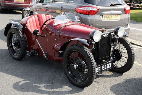A maroon low two-seater with a pointed tail, red seats and wire wheels in a car park. Opens a larger view