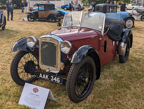 A maroon sports two-seater with black wings, a radiator cowl and wire wheels on grass, a display card in front. Opens a larger view