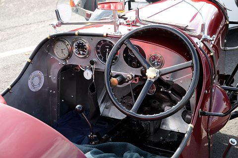 A maroon dashboard with a row of round dials behind a black four-spoke steering wheel. Opens a larger view