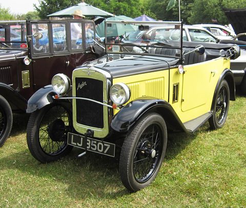 A yellow open tourer with black wings, a folded hood and wire wheels parked on grass among other cars. Opens a larger view