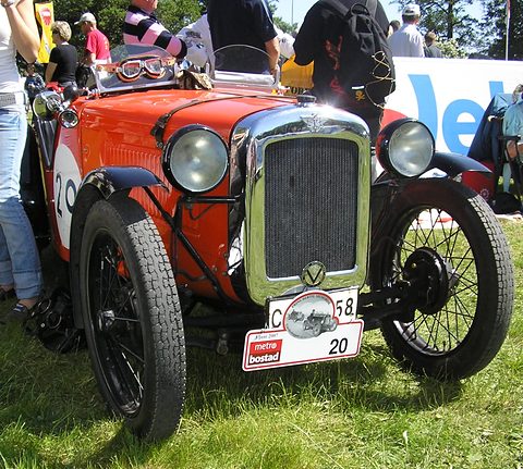 An orange two-seater with a cream radiator, wire wheels and rally plates among people on grass. Opens a larger view