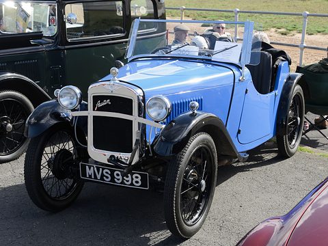 A light blue open two-seater with dark wings and a chrome radiator in a car park. Opens a larger view