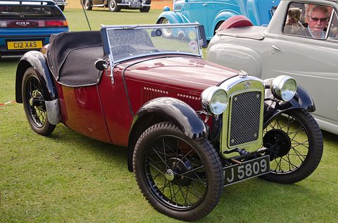 A maroon sports two-seater with black wings, a black hood and wire wheels on grass. Opens a larger view