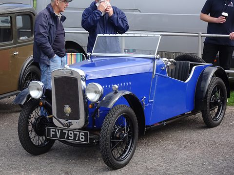 A bright blue open two-seater with black wings and wire wheels in a car park, men standing behind. Opens a larger view