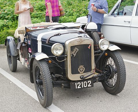 A dark blue open two-seater with a white bonnet stripe, wire wheels and a spare on its side, in a car park. Opens a larger view
