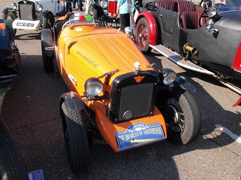 An orange two-seater with a black radiator and black wings parked close among other cars. Opens a larger view