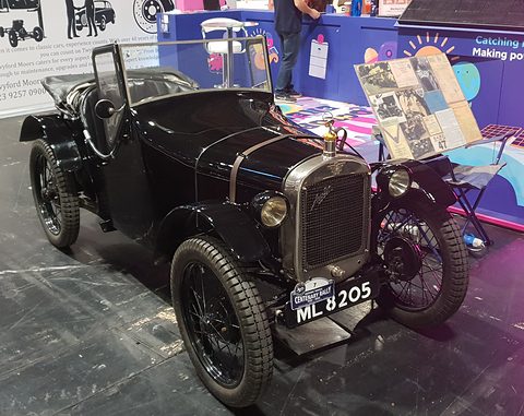A black doorless sports two-seater with wire wheels on a show stand, display boards behind. Opens a larger view