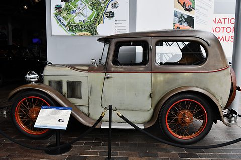 Side view of a weathered grey and brown saloon with red wire wheels and a label in a museum. Opens a larger view
