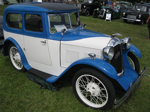 A white saloon with a blue roof and wings and white wire wheels on grass, other cars behind. Opens a larger view