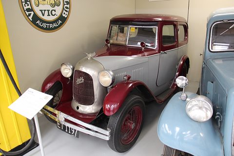 A silver saloon with a maroon roof and wings and a polished radiator cowl in a museum. Opens a larger view