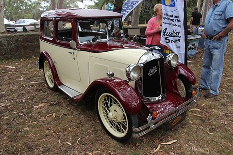 A two-tone cream and maroon saloon with a chrome radiator shell and wire wheels on grass, visitors beside it. Opens a larger view