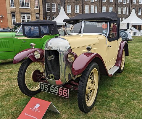 A cream two-seater with maroon wings, a polished radiator cowl and wire wheels on grass, marquees behind.