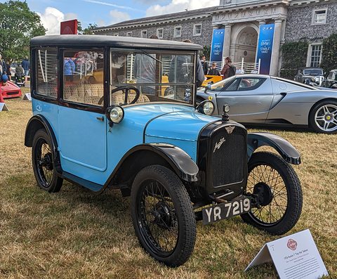 A tall, narrow pale blue saloon with black wings and wire wheels on grass before a grand house. Opens a larger view