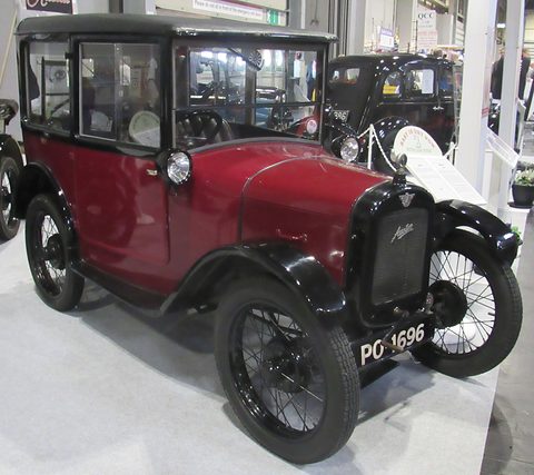 A tall maroon saloon with black roof and wings and wire wheels on a white show floor. Opens a larger view