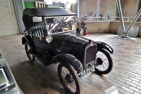 A dark open tourer with its hood up and wire wheels on a brick floor beside a white wall. Opens a larger view
