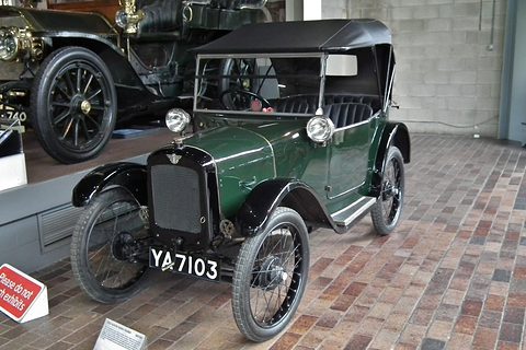 A dark green open tourer with its hood up and wire wheels on a brick floor, other cars beyond. Opens a larger view
