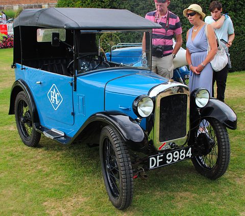 A bright blue open tourer with a black hood, black wings and wire wheels on grass, visitors beside it. Opens a larger view