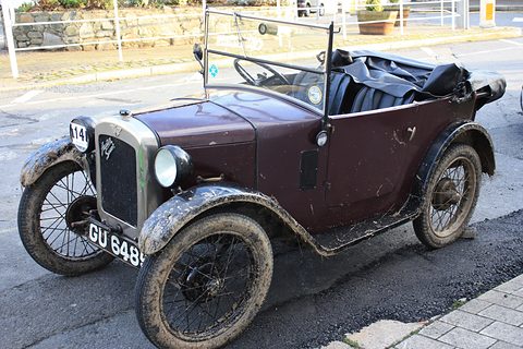 A small maroon open tourer with its hood folded and muddy wheels parked by a kerb. Opens a larger view