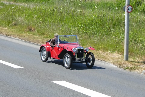 A small red open two-seater driving along a grass-verged road past a 40 speed-limit sign. Opens a larger view