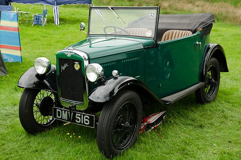 A dark green open two-seater with black wings and wheels on grass, its windscreen up. Opens a larger view