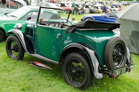 The rear quarter of a green open two-seater with a folded black hood and a spare wheel on its tail. Opens a larger view