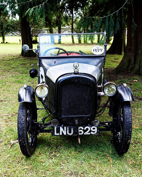 Head-on view of a dark two-seat sports car with a black radiator cowl, cycle wings and a tall windscreen. Opens a larger view