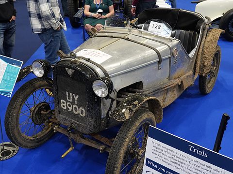 A muddy grey sports two-seater numbered 128 on a blue show floor beside a sign reading Trials. Opens a larger view