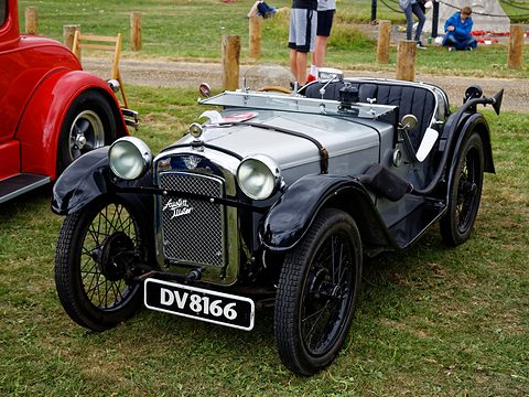 A silver doorless sports two-seater with black wings and wire wheels on a village green. Opens a larger view