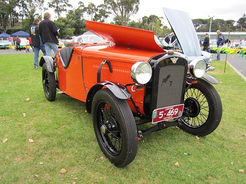 An orange doorless two-seater with its bonnet sides raised on grass, visitors behind. Opens a larger view