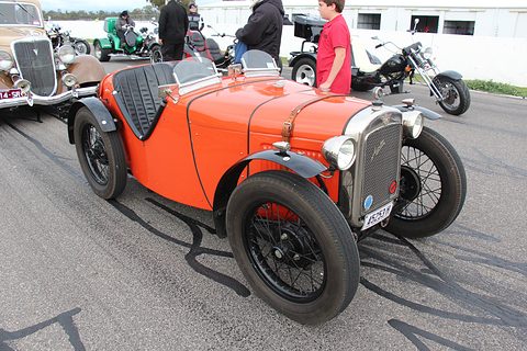 An orange doorless sports two-seater with black wings and wire wheels in a paddock, people behind. Opens a larger view