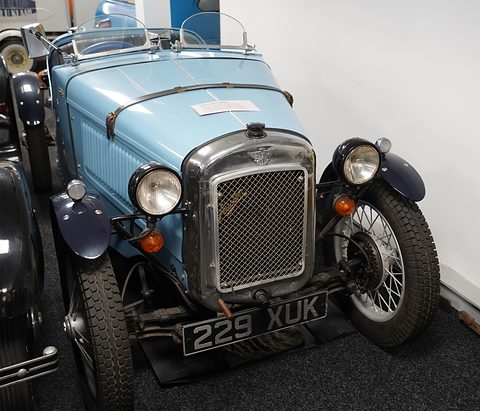 A pale blue doorless sports two-seater with black wings and wire wheels on a dark museum floor. Opens a larger view