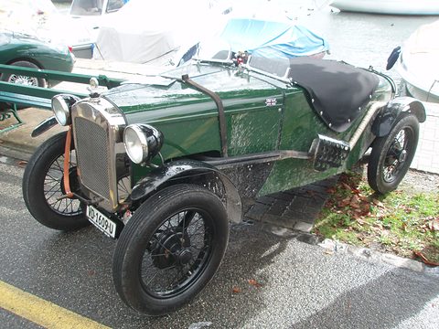 A dark green doorless sports two-seater with a tonneau cover and wire wheels on a lakeside quay. Opens a larger view