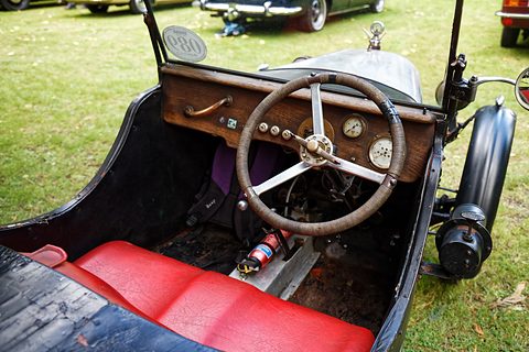 Looking into a doorless sports car over red seats to a wooden dashboard with dials and a large steering wheel. Opens a larger view