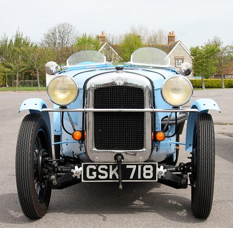 Head-on view of a pale blue two-seater with a tall radiator, large headlamps and twin aero screens. Opens a larger view