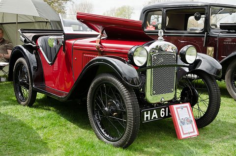 A low red open two-seater with black wings, a chrome mesh radiator, wire wheels and a placard on the grass. Opens a larger view
