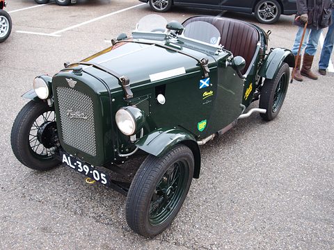 A dark green doorless sports two-seater with wire wheels and a Dutch plate on paving. Opens a larger view