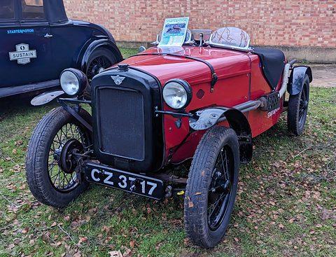 A red doorless sports two-seater with black wings, a black radiator and wire wheels on grass before a brick wall. Opens a larger view